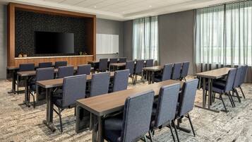 Une salle de conférence équipée d’un grand téléviseur à écran plat, de plusieurs tables et chaises, et dotée d’un sol recouvert de moquette.