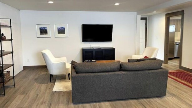 Un salon moderne avec un canapé gris, deux fauteuils blancs, un téléviseur à écran plat et un plancher en bois.
