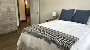 Une chambre à coucher comprenant un lit, une table de chevet, une lampe, une armoire et une porte ouverte donnant sur une autre pièce.
