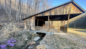 A rustic wooden structure with a covered porch, a small stream, and surrounding natural landscape.