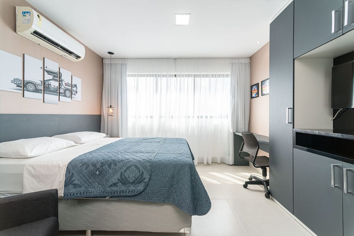 A hotel room with a bed, a desk, a chair, an air conditioning unit, and a television.