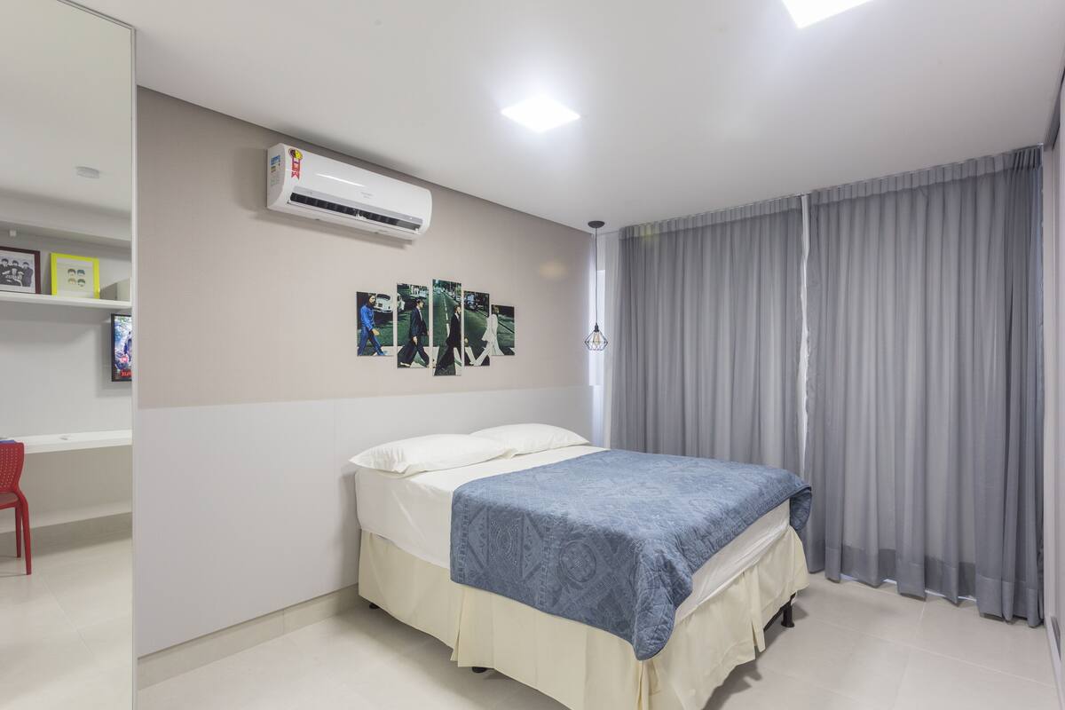 A bedroom with a bed, an air conditioner, a desk with a chair, and a window with curtains.