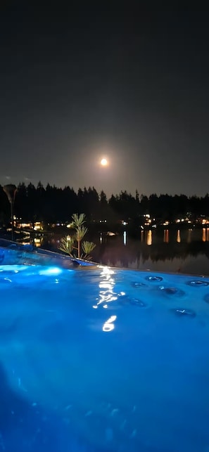 A nighttime pool area with a moonlit sky, a calm water surface reflecting lights, and a distant tree line.