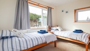 A room with two single beds, a window with curtains, a small table with a lamp, and a view of greenery outside. - Brookfield (Bude)