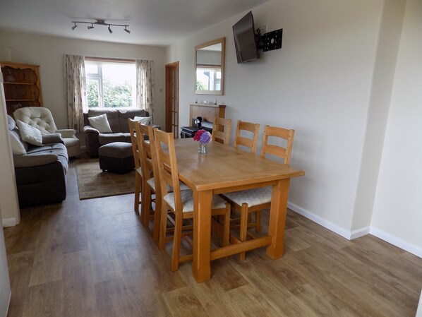 A spacious living room with a wooden dining table, comfortable seating, a television mounted on the wall, and a view through the window. - Brookfield (Bude)