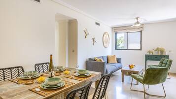 Un coin repas avec une table en bois dressée pour quatre personnes, entourée de chaises en osier. Un canapé et des chaises vertes se trouvent à l’arrière-plan.