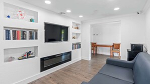 A modern living room with a flat-screen TV, bookshelves, a fireplace, a dining area, and a gray sofa. - Travelodge by Wyndham Kissimmee/Orlando (Kissimmee)