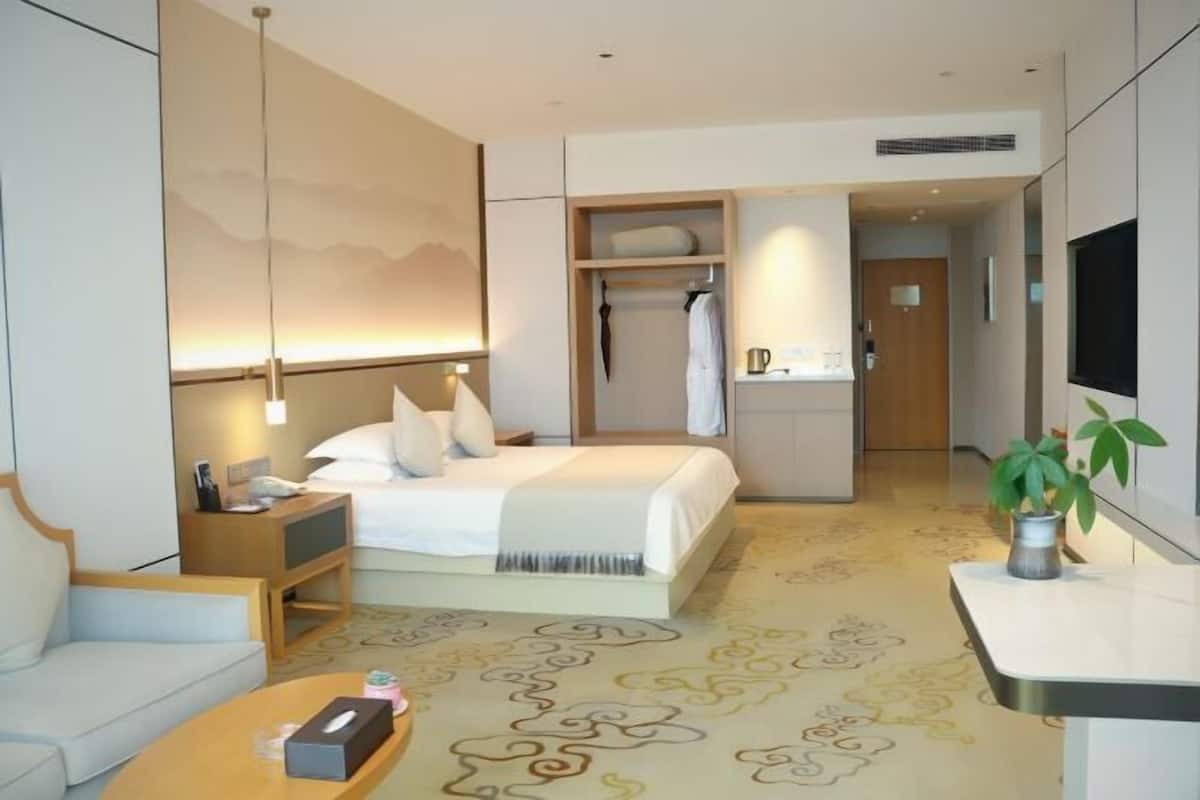 A modern hotel room with a bed, bedside table, and a small seating area. There is a TV mounted on the wall, a wardrobe, and a bathroom visible through an open door.