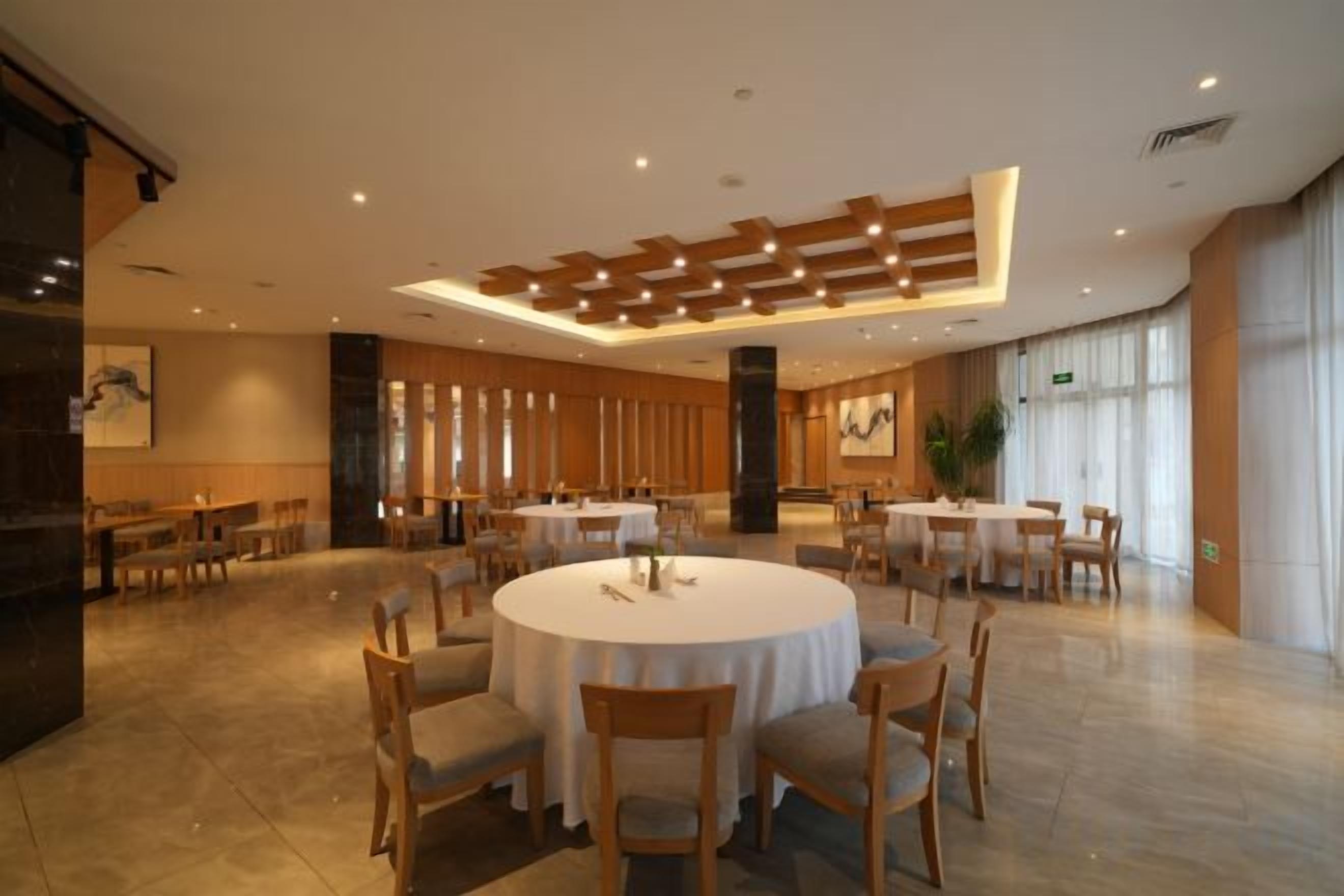 A spacious hotel dining area with round tables and chairs, large windows, and a modern ceiling design.