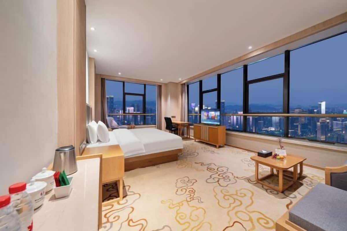 A modern hotel room with a large bed, a desk, a chair, and a view of the cityscape through large windows.