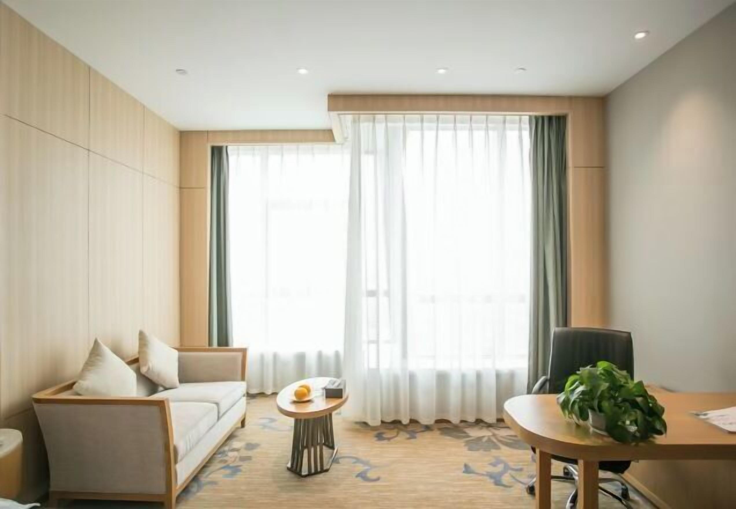A modern hotel room with a sofa, a small round table with a fruit bowl, a chair, and a potted plant.