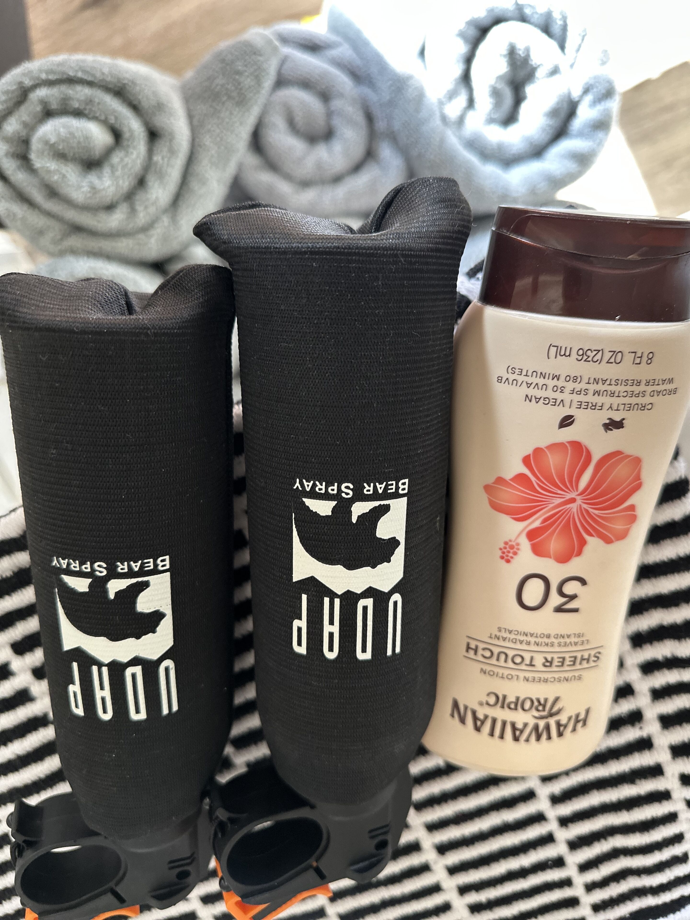 A pair of black socks with a bear design and the text "BEAR SPAY" on them, placed next to a bottle of sunscreen and a rolled towel.
