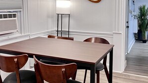 Ein Konferenzraum mit einem rechteckigen Tisch, vier Holzstühlen, einer Stehlampe und einem runden Spiegel an der Wand.