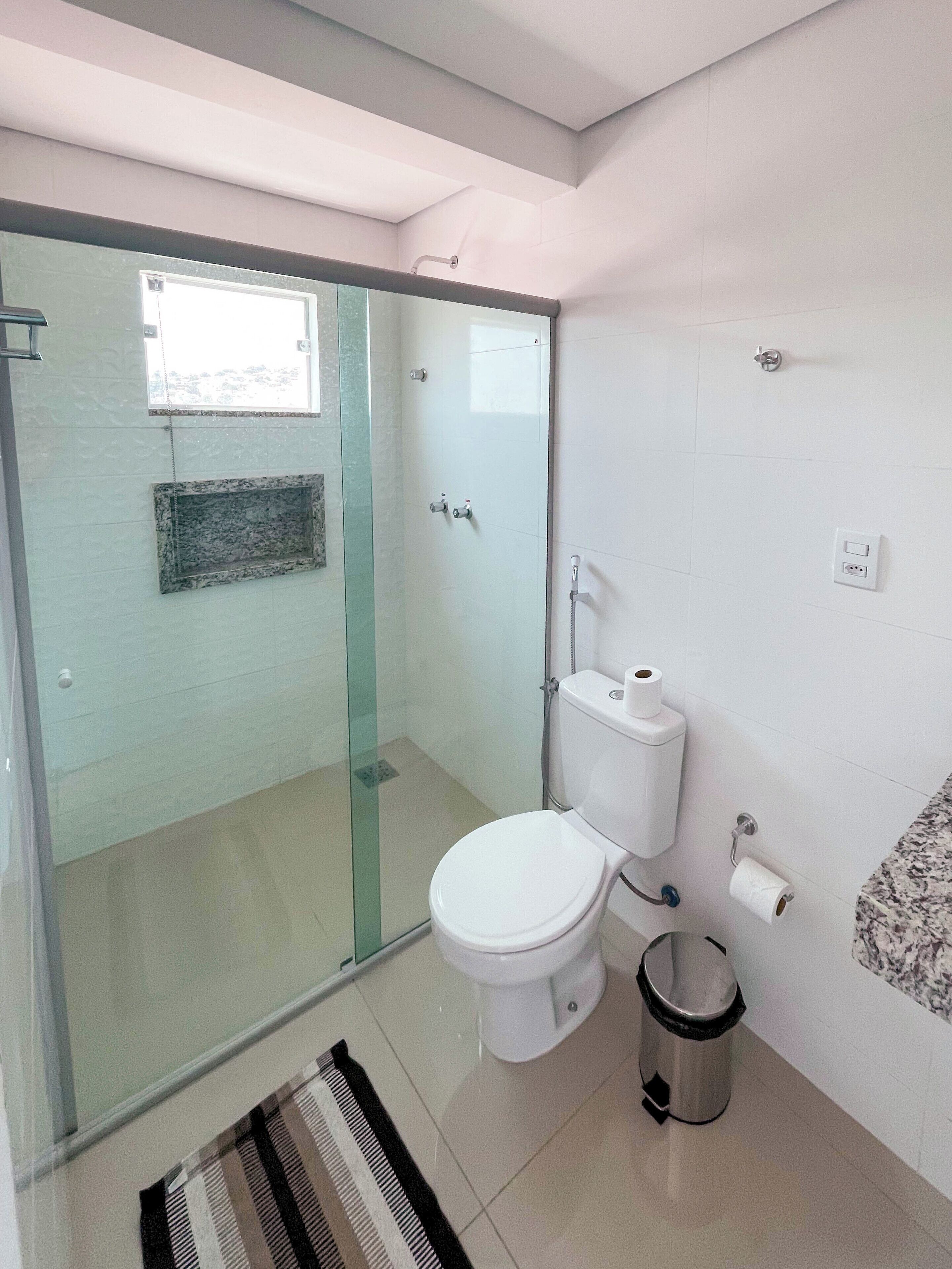 Baño con cabina de ducha de vidrio, inodoro, cubo de basura y perchero para toallas.