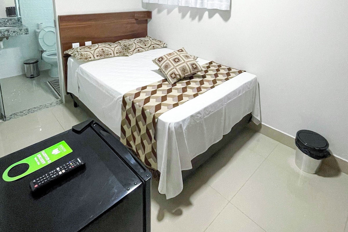 Habitación de hotel con una cama, una mesita con un control remoto, un cesto de basura y una ventana con cortinas.
