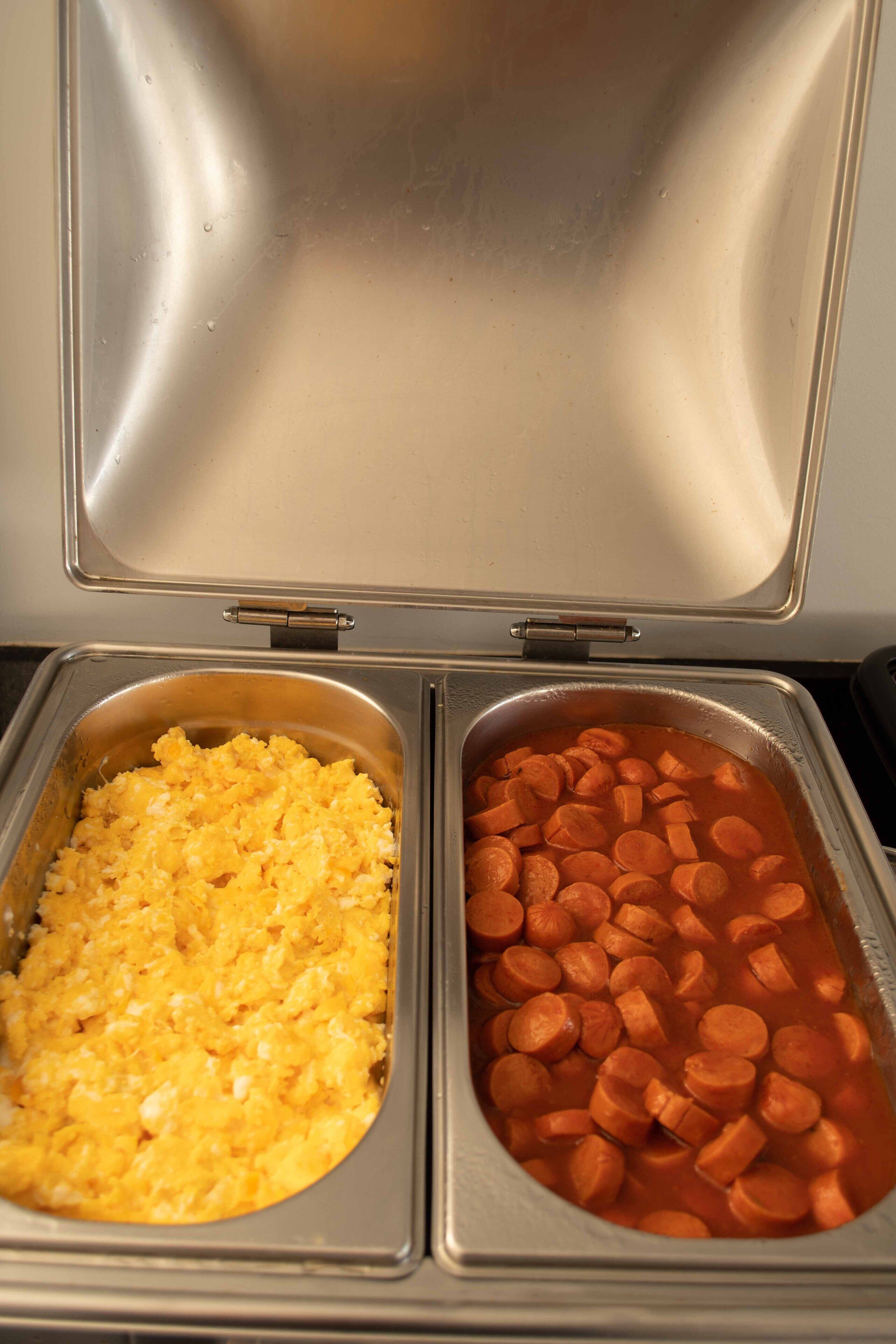 Un recipiente de acero inoxidable para servir alimentos, con dos compartimentos, uno con huevos revueltos y el otro con zanahorias en rodajas.