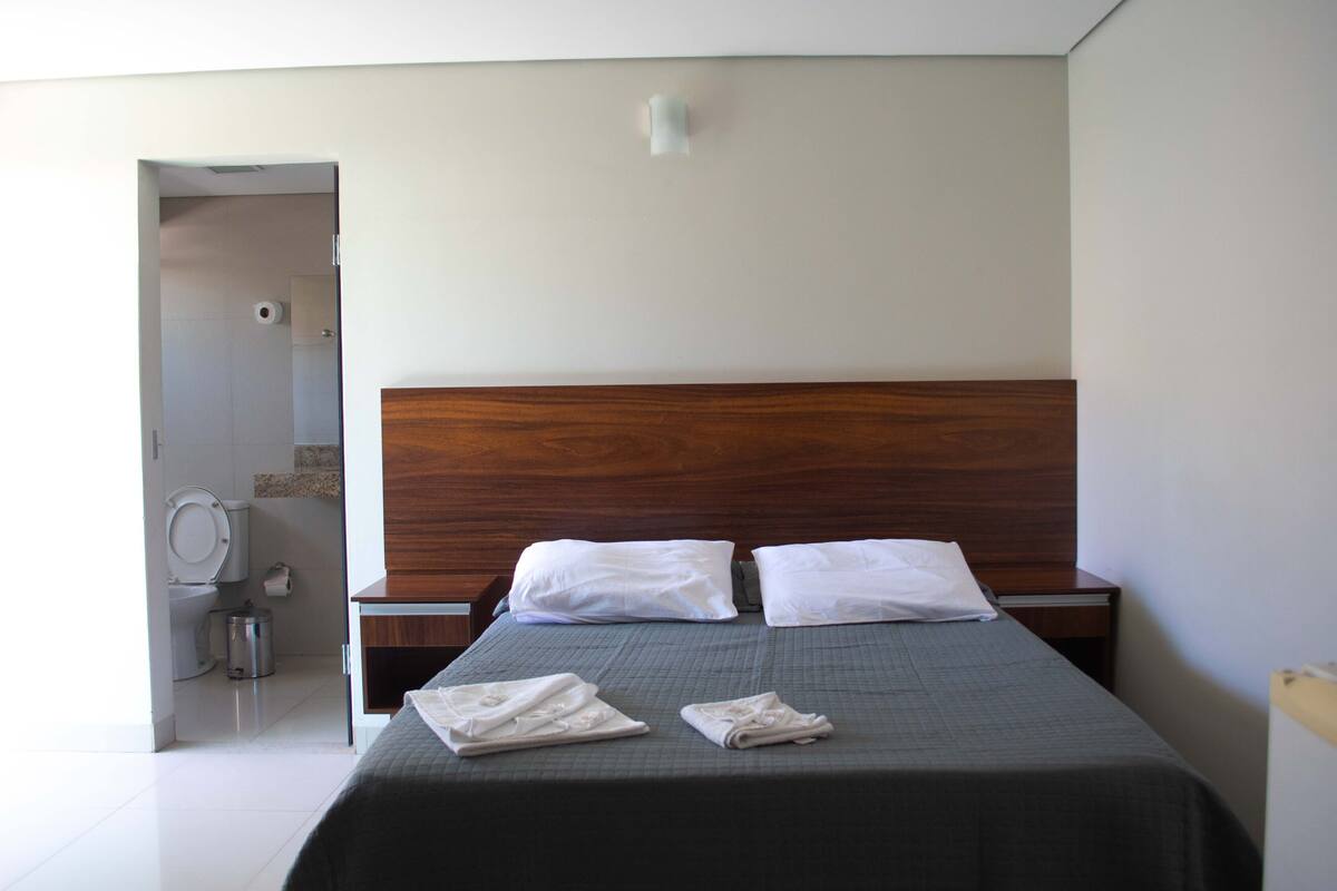Habitación de hotel con cabecera de madera, dos almohadas blancas, una colcha gris y dos toallas dobladas sobre la cama.