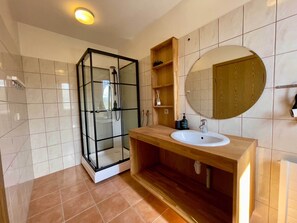 A bathroom with a glass-enclosed shower, a wooden vanity with a sink, and a round mirror. - Matkráin Apartments (Hveragerdi)