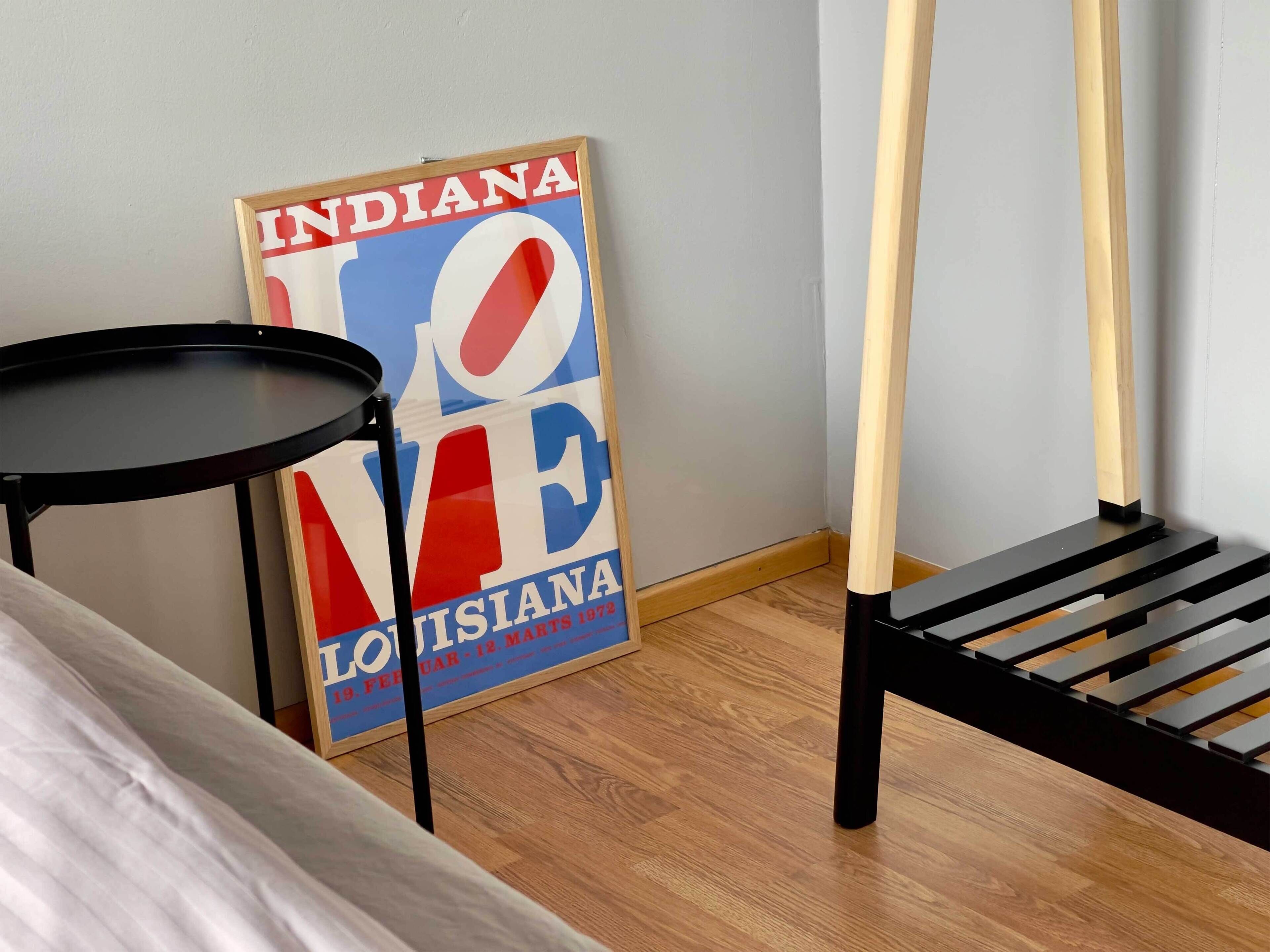 A room with a bed, a small round table, a black chair, and a poster on the wall.