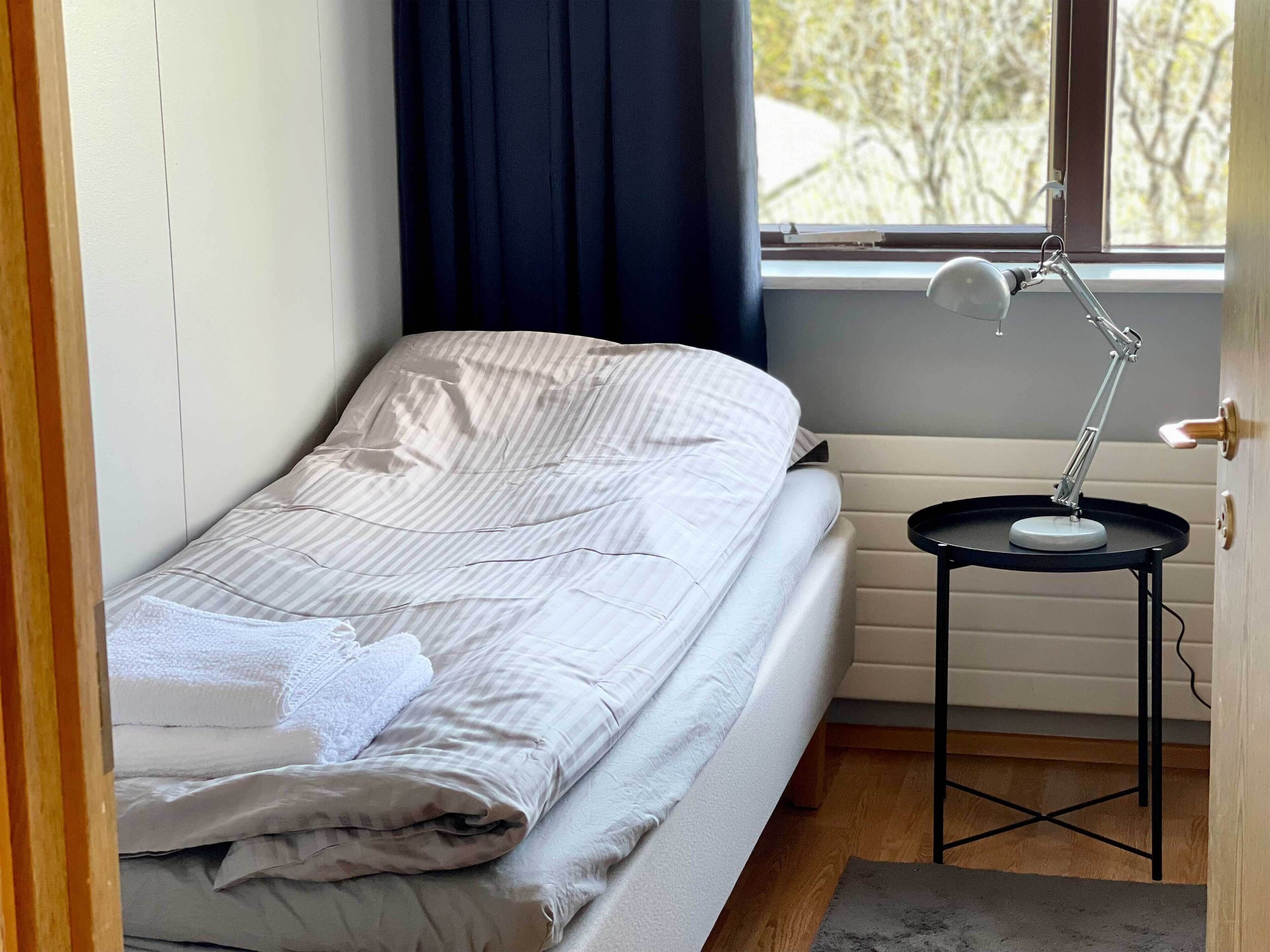 Una cama pequeña y ordenada con una manta a rayas, una mesita de noche con una lámpara, y una ventana que da a vistas de árboles.