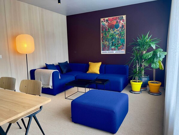 A modern living room with a blue sofa, a coffee table, a wooden dining table, and potted plants. - Matkráin Apartments (Hveragerdi)