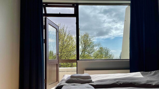 Habitación de hotel con cama, ventana con cortinas y vista a árboles y cielo nublado.