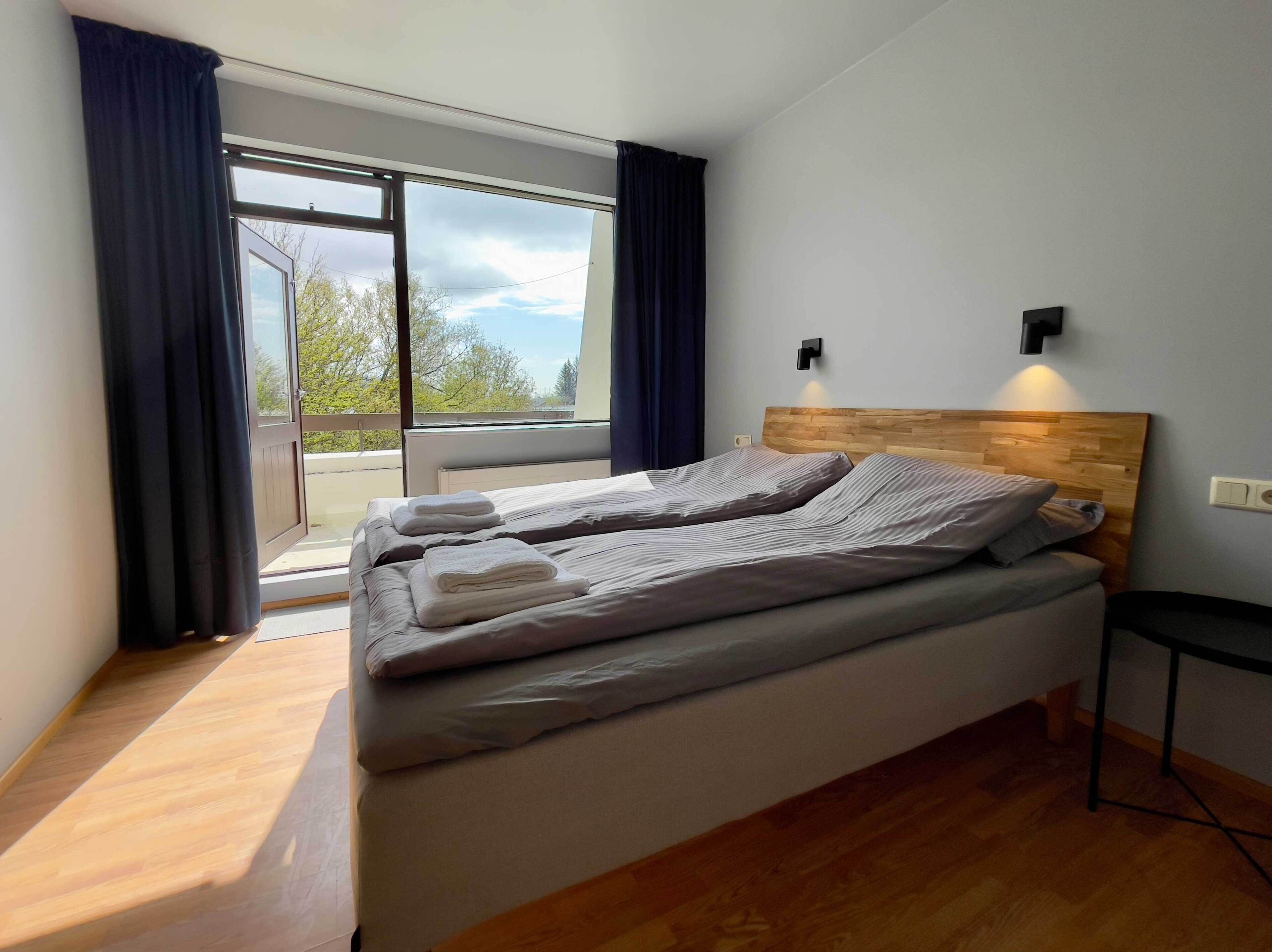 A bedroom with a bed, a nightstand, a wall-mounted light, and a view through a large window.