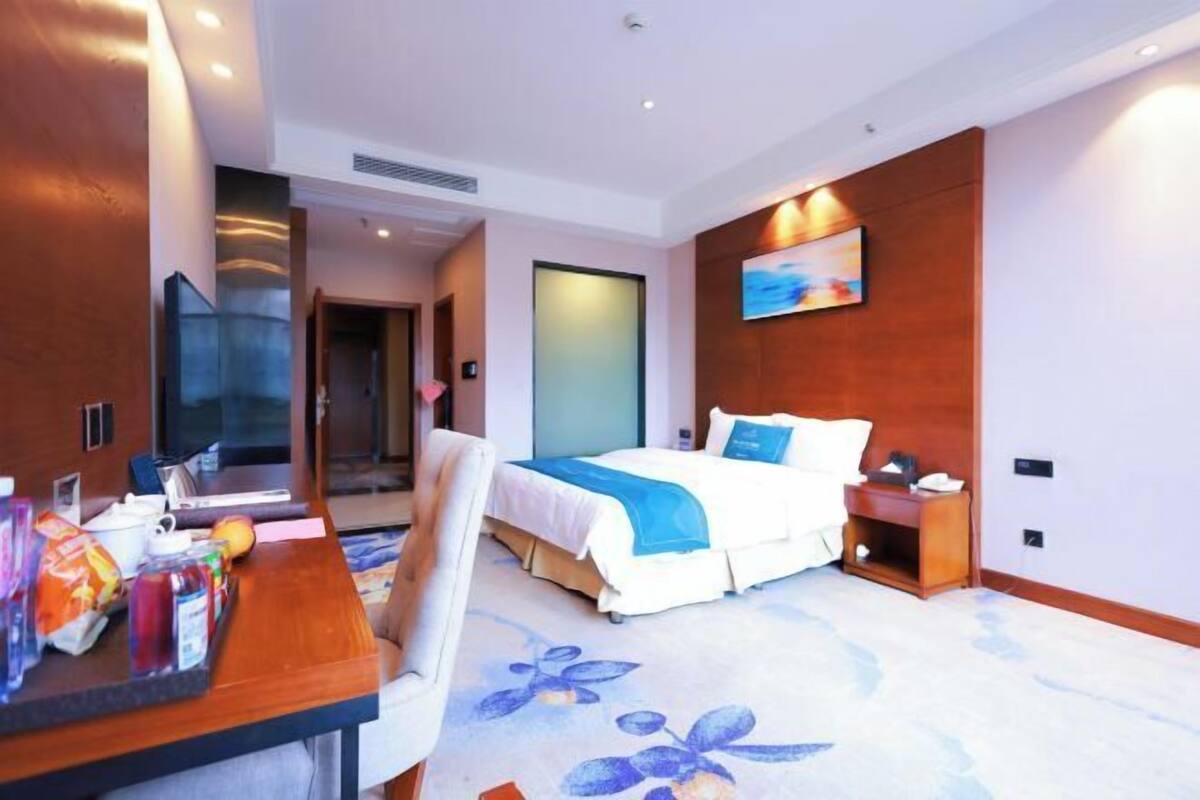A hotel room with a bed, bedside table, chair, and a desk with snacks and a laptop.