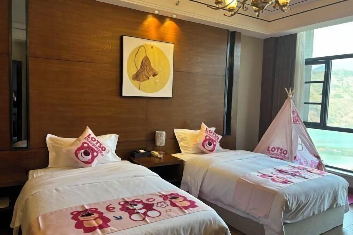 A hotel room with two beds, each with a pink and white LOTSO-themed bedspread. There is a painting on the wall above the beds and a window with a view of the outdoors.