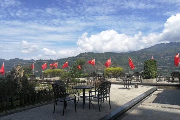 An outdoor seating area with tables and chairs, red flags, and a mountainous backdrop. - Huamei Hotel (Suijiang)