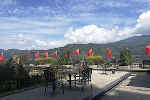 Un área de estar al aire libre con mesas y sillas, banderas rojas y montañas al fondo.