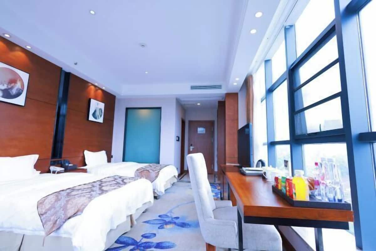A hotel room with two beds, a desk with a chair, a TV, and large windows.