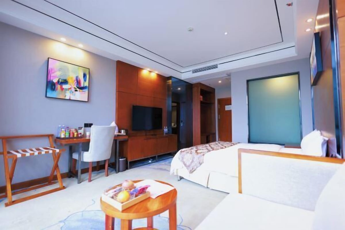 A hotel room with a bed, a desk, a TV, a chair, and a small table with fruits.