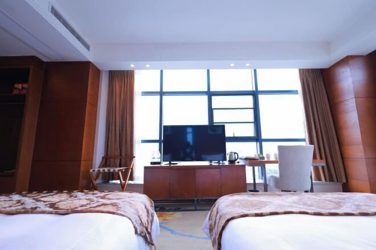 A hotel room with two beds, a desk, a television, and a large window.