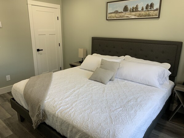 Un dormitorio con cama, mesita de noche, lámpara, puerta y un cuadro en la pared.