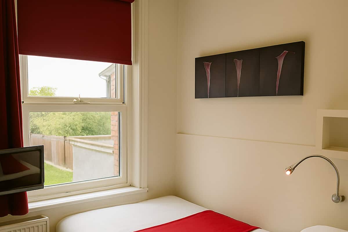 A neatly made bed with white and red bedding, a television on a stand, a window with red curtains, and a wall-mounted artwork.