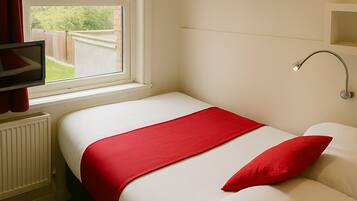 Una cama bien hecha con ropa de cama blanca y roja, un televisor sobre un mueble, una ventana con cortinas rojas y una obra de arte colgada en la pared.