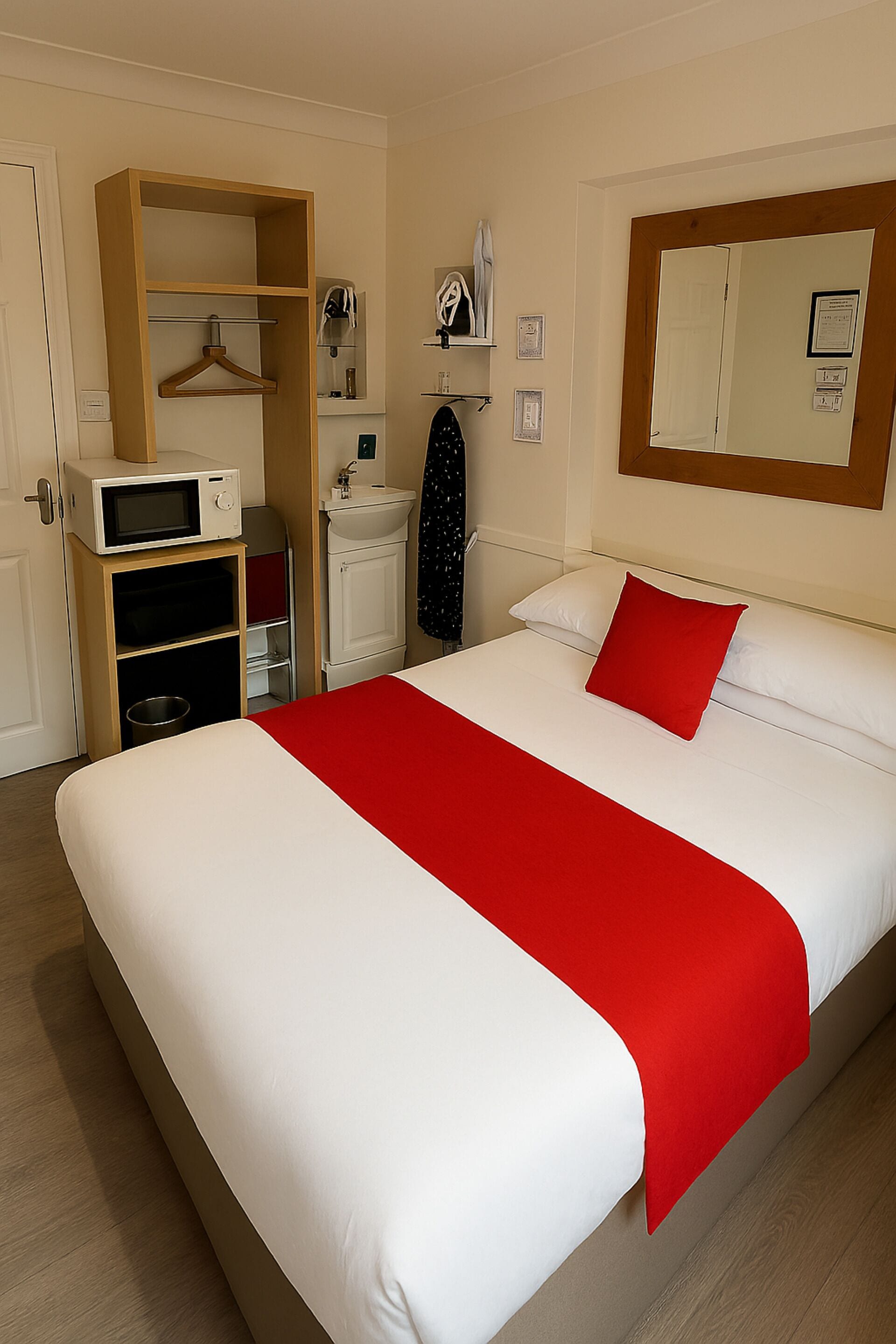 A hotel room with a large bed, a red throw blanket, a wooden wardrobe, a microwave, a sink, and a mirror on the wall.