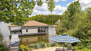 Ein Hausboot an einem See, mit einer Terrasse, Gartenmöbeln und einem Sonnenschirm.