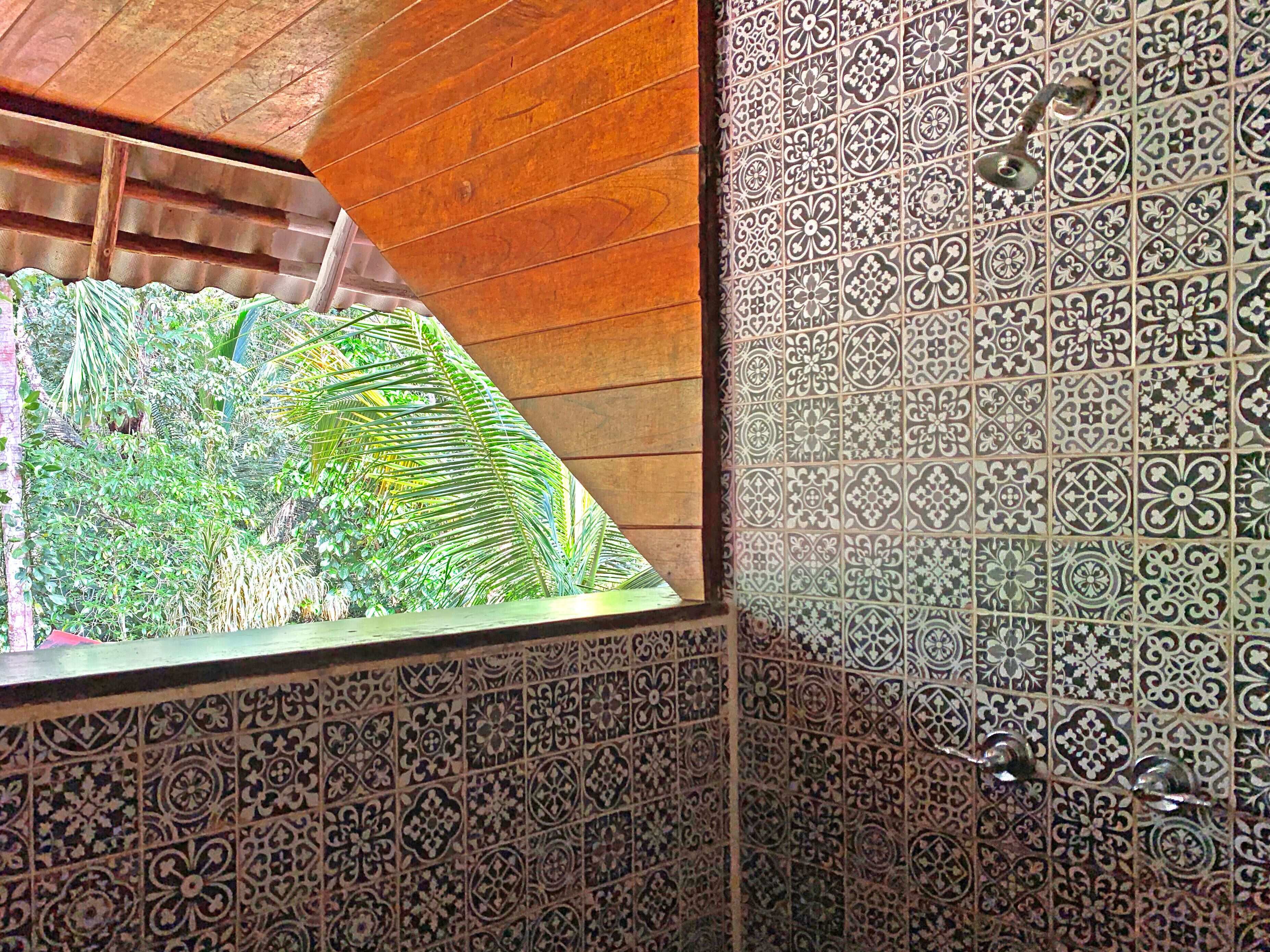 A room with a sloped wooden ceiling, a window with a view of lush greenery, and a wall with intricate tile patterns.