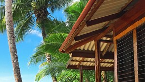 Eine Holzhütte mit überdachter Veranda mit Blick auf einen Strand mit Palmen und wolkenlosem, blauem Himmel.
