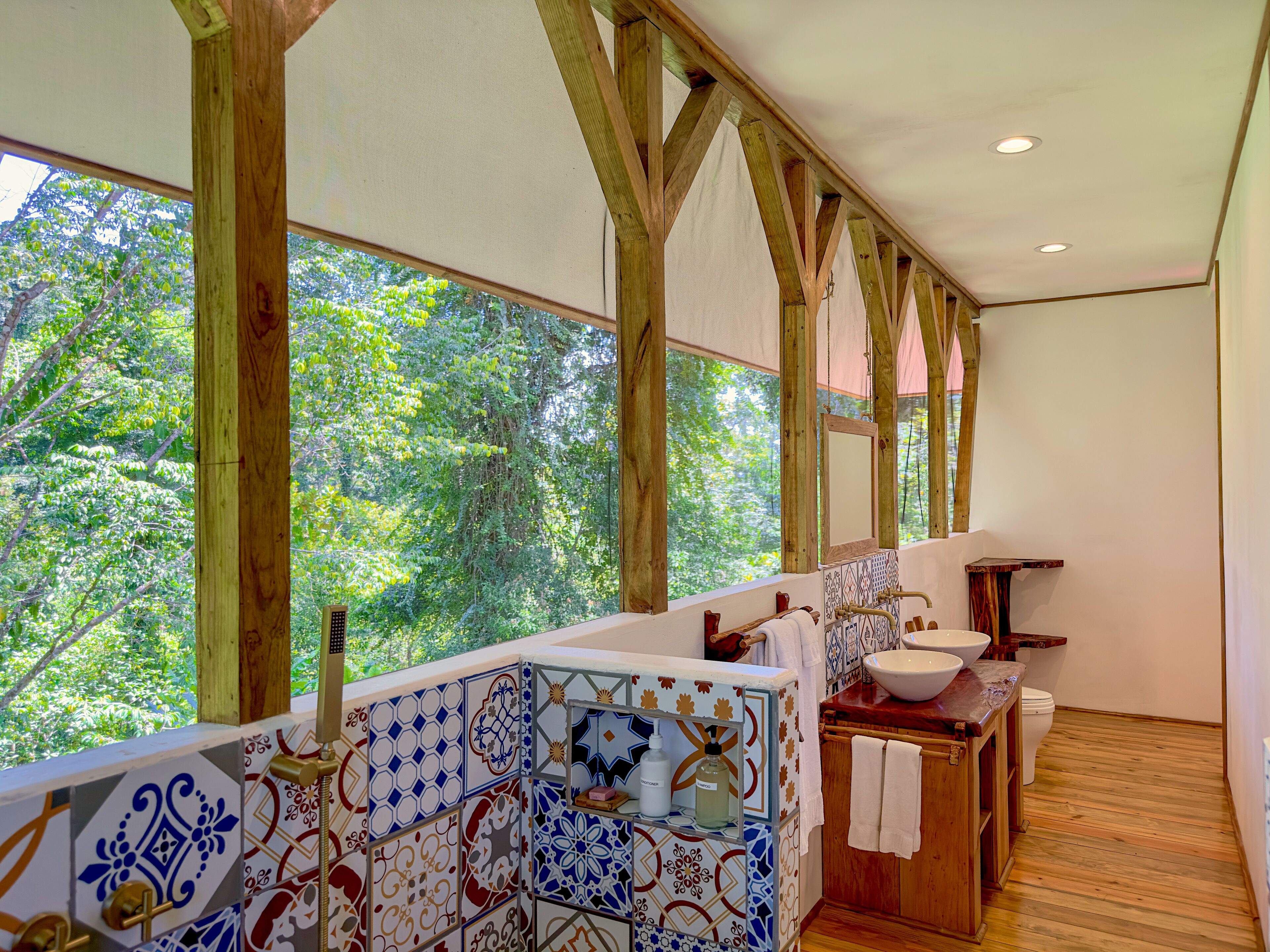 A wooden balcony with a view of lush greenery, a tiled wall with a sink and mirror, and a wooden floor.