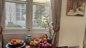 Een kamer met een raam, een tafel met fruit, een vaas met bloemen en een schilderij aan de muur.