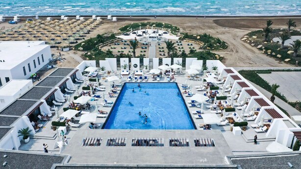 A resort with a central swimming pool surrounded by sun loungers and umbrellas, a beach area with umbrellas and lounge chairs, and a view of the sea.