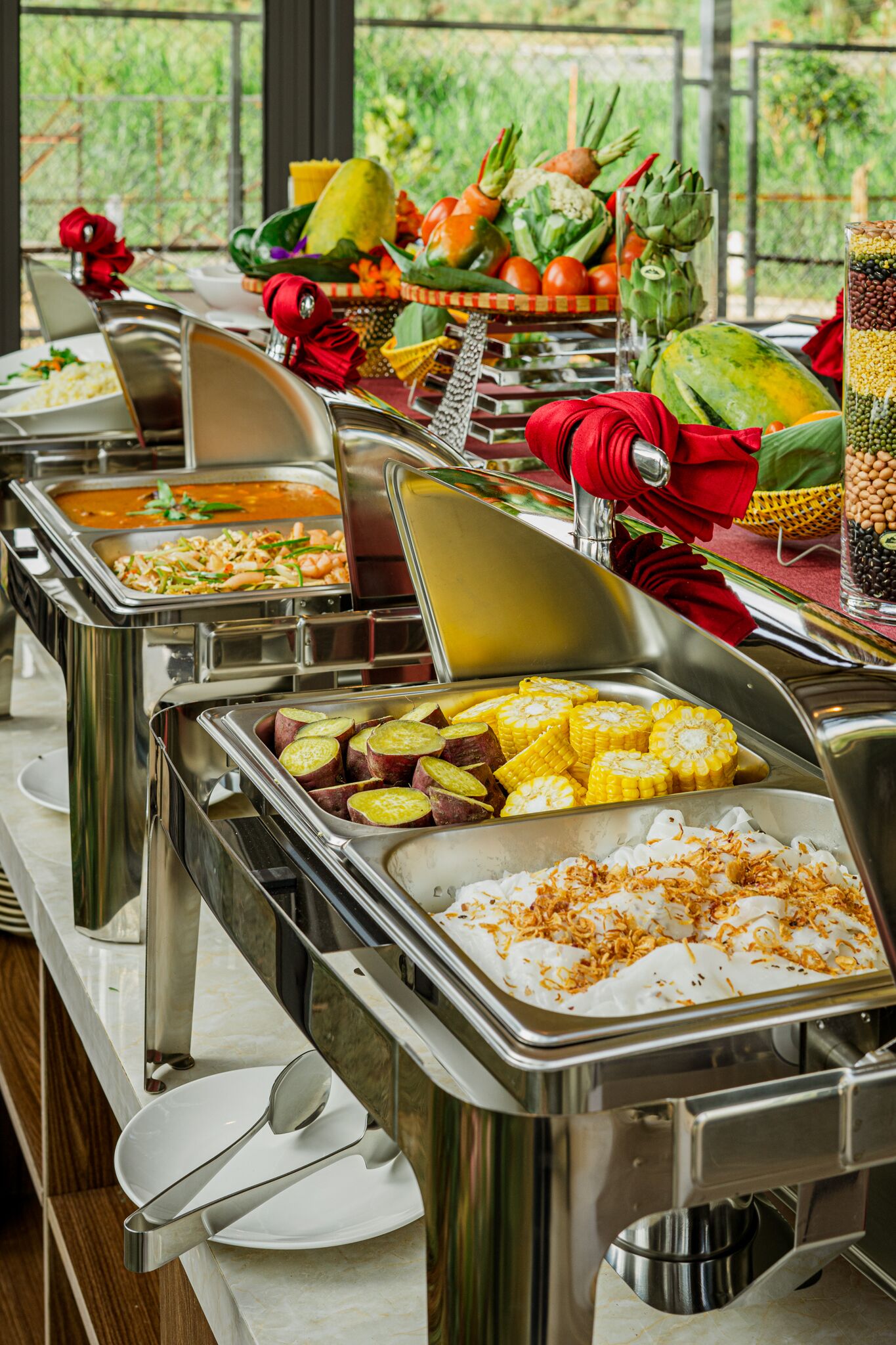 A buffet with various food items and a view of greenery outside.