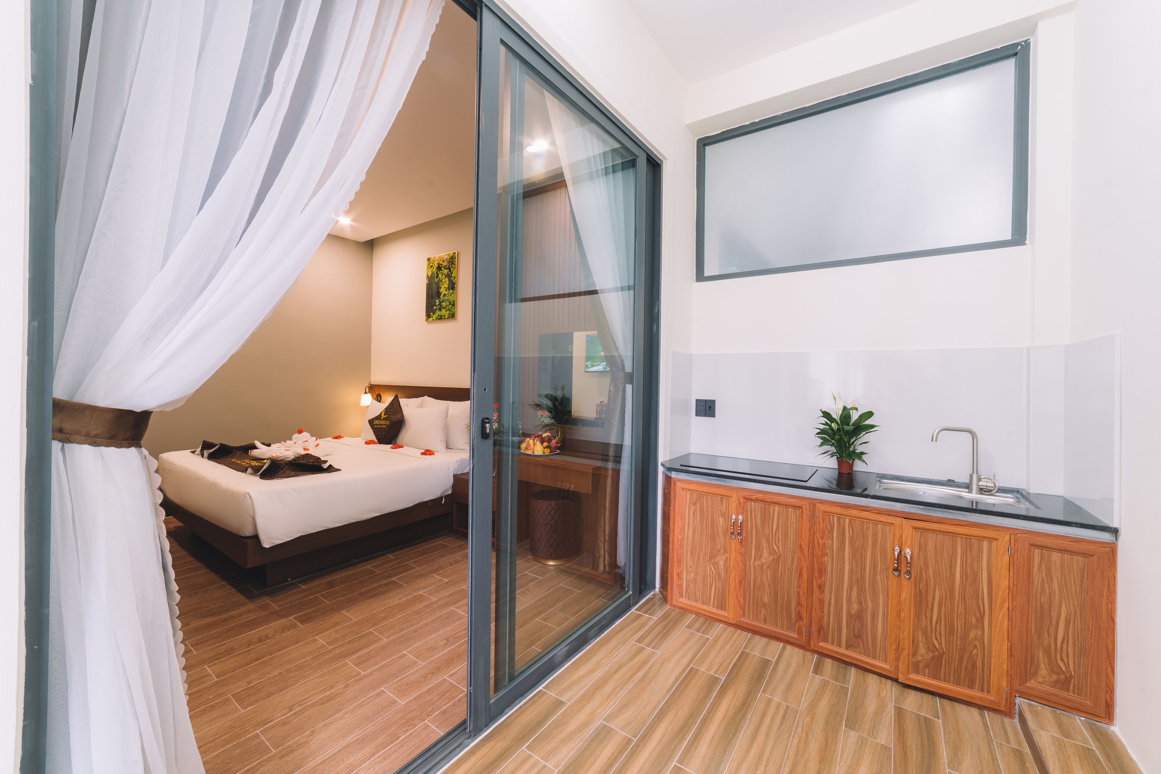 A bedroom with a bed, a sliding glass door, a wooden cabinet, and a small kitchen area with a sink and a potted plant.