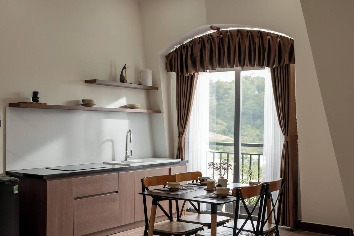 A compact kitchen with a sink, wooden cabinets, a black refrigerator, and a dining table with chairs. A window with curtains provides a view of greenery outside.