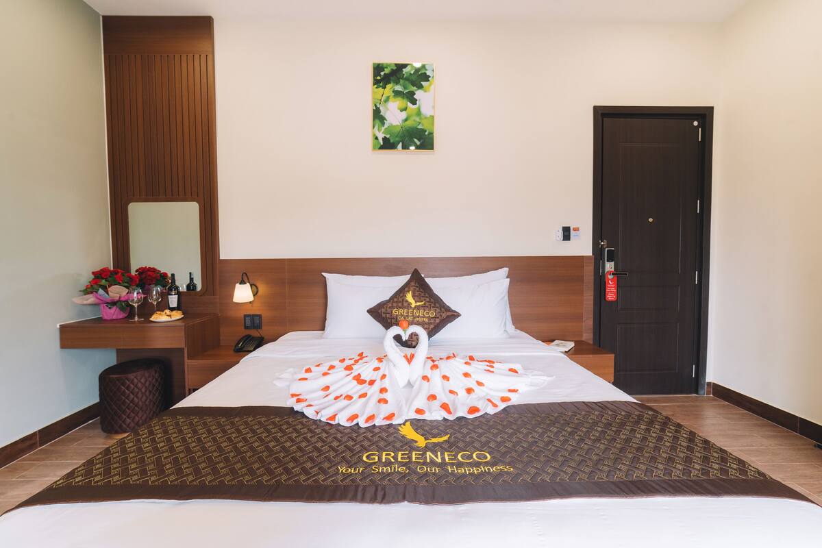 A hotel room with a neatly made bed, a wooden headboard, a small table with a vase of flowers, and a door.