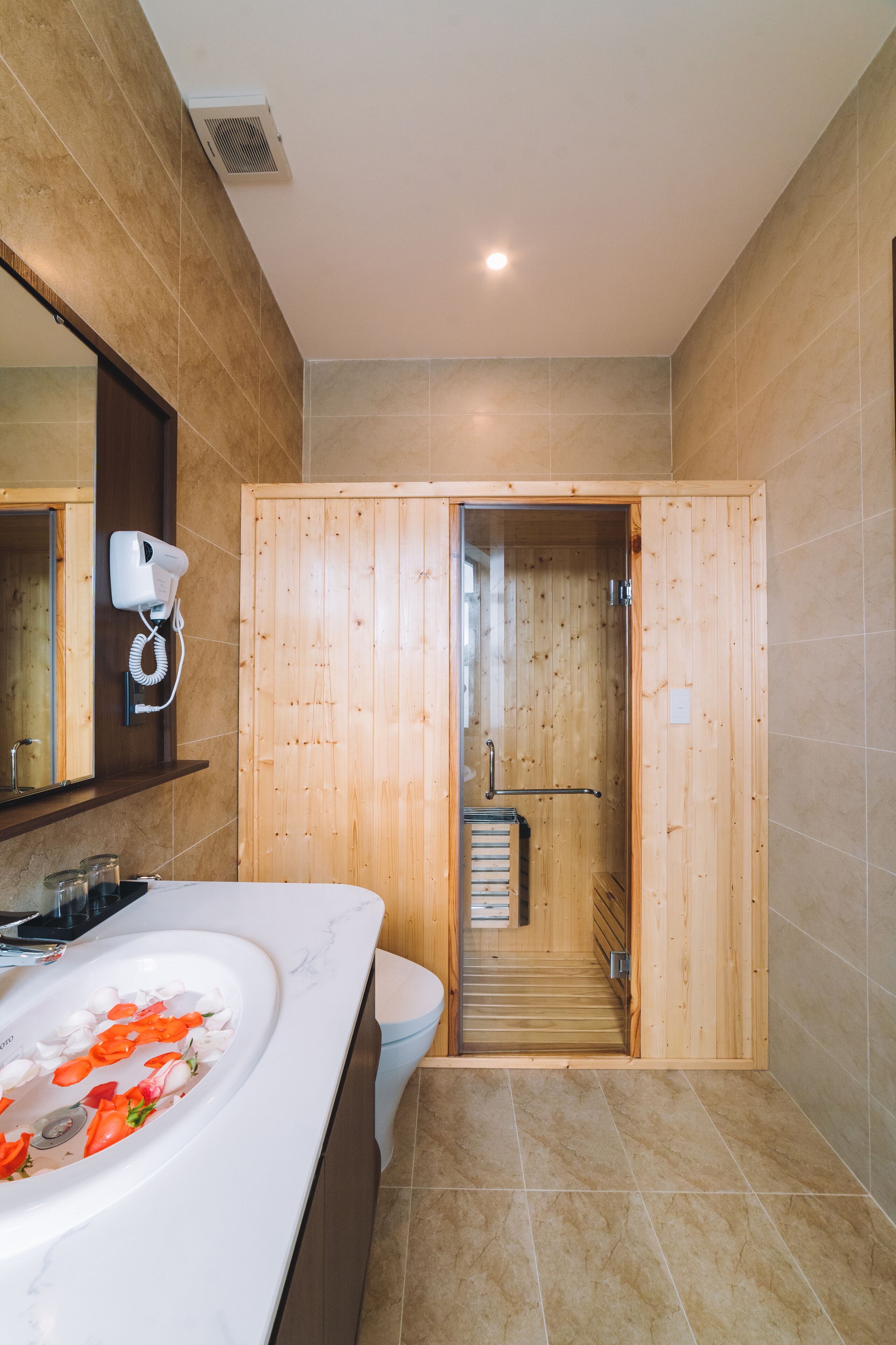 Un baño moderno con una puerta de sauna de madera, un lavamanos blanco adornado con pétalos de rosa y un secador de pelo montado en la pared.