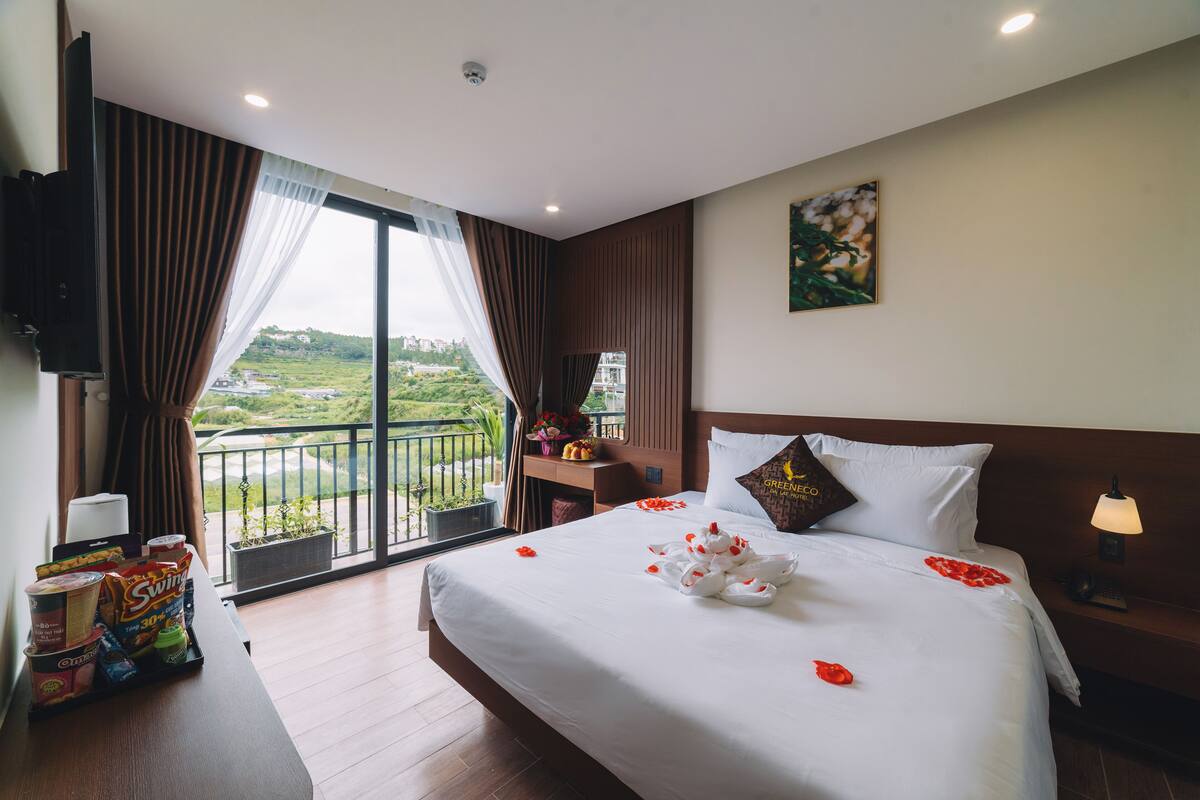 A hotel room with a large bed, a TV, a bedside table with a lamp, and a view of a balcony with greenery.